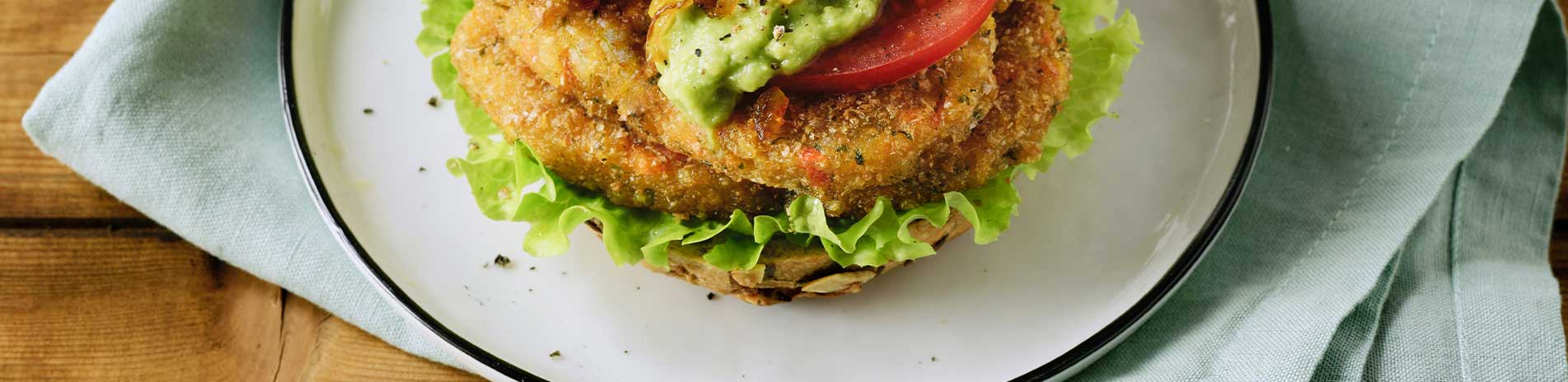 Veggie Love Süßkartoffel Patties mit Tomate, Salat und Guacamole in einem Brötchen liegen auf einem Teller