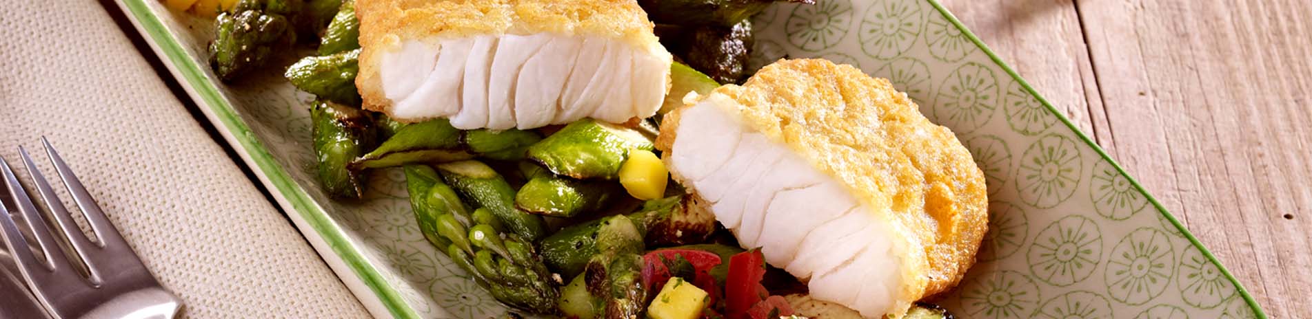 Backfisch-Happen auf grünem Spargel, Mais und roter Parika in einer Schale, daneben eine Gabel und eine Stoffserviette