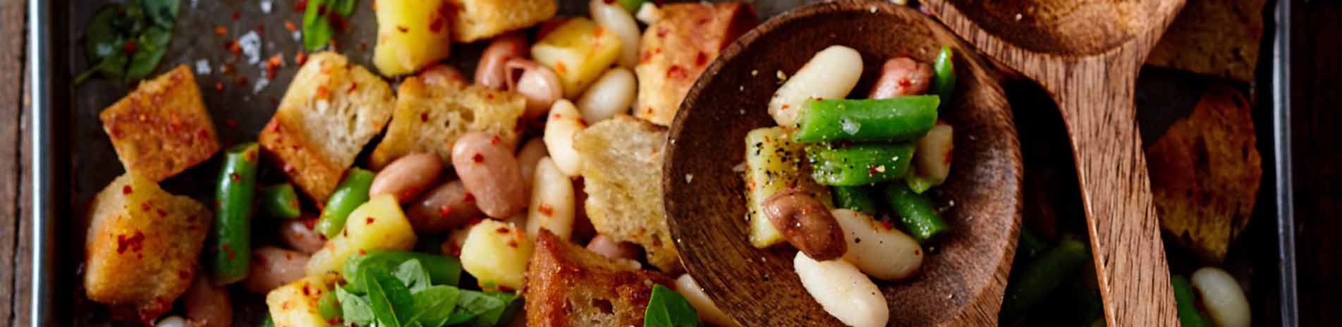Verschiedene Bohnen und geröstete Brotwürfel auf einem Blech, sowie zwei Holzlöffel
